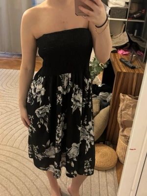 Svart axelbandslös blommig klänning - Säljer en svart axelbandslös klänning med vita blommor. Klänningen har smock upptill för en bekväm passform och ett flowy kjolparti som slutar strax ovanför knäna. Perfekt för varma dagar och enkel att styla med accessoarer.