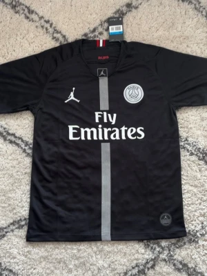Svart PSG x Jordan matchtröja - Svart PSG matchtröja i samarbete med Jordan, med Fly Emirates-tryck på bröstet och PSG-logga. Tröjan har en grå vertikal rand i mitten och klassisk rund hals. Tillverkad i lätt och ventilerande material, perfekt för fotbollsträning.