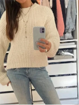 Krämvit stickad tröja med rund hals - Mysig krämvit stickad tröja med rund hals och breda ärmar. Tröjan har en grovstickad struktur och avslappnad passform, perfekt för lager-på-lager. Passar till jeans eller kjol för en chill look.