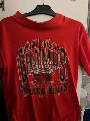Röd Chicago Bulls t-shirt - Strl L/M —->Säljer en röd Chicago Bulls t-shirt med stort tryck på framsidan som firar Eastern Conference Champs och 25th Anniversary. Klassisk rund hals och korta ärmar, perfekt för dig som gillar basket och streetwear. 