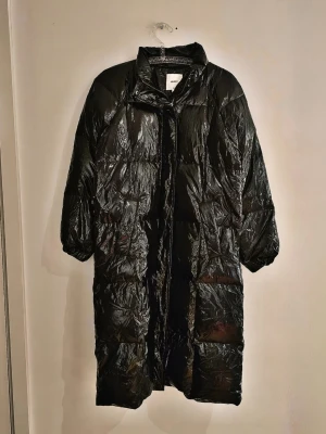 Svart lång dunjacka från Samsøe Samsøe - Snygg svart lång dunjacka från OBJECT med glansig finish och hög krage. Jackan har raka ärmar, dold dragkedja och tryckknappar framtill samt två sidofickor. Perfekt för kalla vinterdagar och ger en cool, modern look.