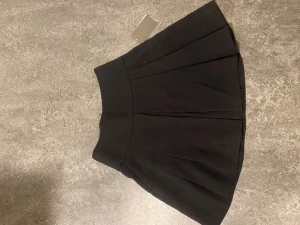 Svart kortkjol från Zara med dragkedja - Snygg svart kortkjol från Zara med markerad midja och veckad design. Kjolen har en synlig dragkedja bak och är perfekt för en trendig look. Tillverkad i ett mjukt material som ger en skön passform.Köpt för 300kr 