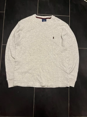 Grå långärmad tröja Polo Ralph Lauren - Mysig grå långärmad loungewear-tröja från Polo Ralph Lauren. Tillverkad i 100% bomull med en diskret broderad logga på bröstet. Tröjan har rund halsringning och ribbade muddar, perfekt för chill dagar hemma.