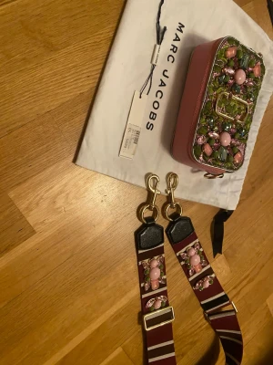 Marc Jacobs axelväska med pärldekor - Axelväska från Marc Jacobs i rosa med rektangulär form och bred, justerbar axelrem. Väskan är dekorerad med pärlor och stenar i rosa och grönt på framsidan samt har guldfärgade metalldetaljer. Märkets logga syns framtill. Materialet är en mix av läder och textil.