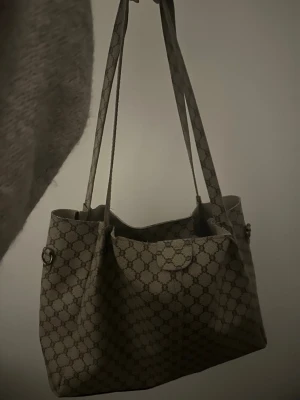  axelväska med monogrammönster - Snygg axelväska i beige och brunt med klassiskt monogram över hela väskan. Väskan har dubbla axelband och silverfärgade metalldetaljer. Tillverkad i canvas med läderdetaljer. Perfekt för dig som gillar lyxiga accessoarer med ikonisk design.