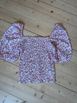 Blommig topp med puffärm från ONLY - Supersöt blommig topp från ONLY med smockad kropp och stora puffärmar. Toppen har en fyrkantig urringning och är i rosa, vit och gul färgskala. Perfekt för dig som gillar romantisk och trendig stil.