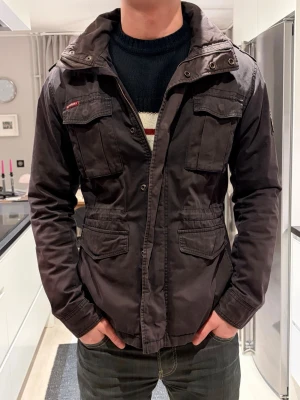 Mörkbrun jacka från Superdry - Snygg mörkbrun jacka från Superdry med fyra stora fickor framtill, hög krage och detaljer som axelklaffar och patch med brittisk flagga på ärmen. Tillverkad i slitstarkt bomullsmaterial och stängs med dragkedja och knappar. Perfekt för dig som gillar utility-stil.