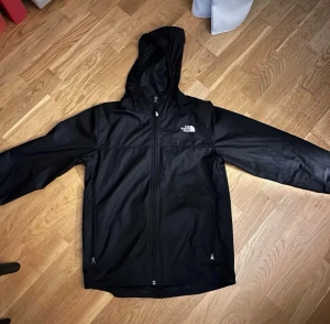 Svart vindjacka från The North Face - Snygg svart vindjacka från The North Face med huva och hel dragkedja framtill. Jackan har två fickor med dragkedja och logga på bröstet. Perfekt för blåsiga dagar och enkel att matcha med olika outfits. Storlek XL i barn passar som en XS, 10/10 skick nästan aldrig använd 
