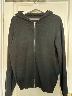 Axel luxe zip - Mysig svart stickad zip med dragkedja framtill. Tröjan har långa ärmar, ribbade muddar och en stor huva. Perfekt för lager på lager och chill dagar. Enkel och stilren design utan synligt märke.