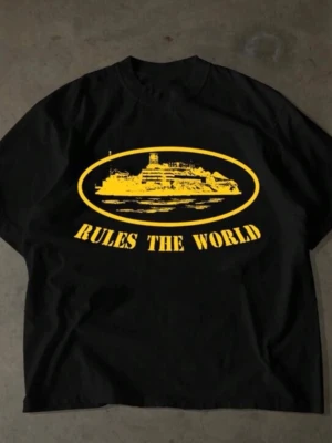 Svart t-shirt med gul print - Cool svart t-shirt med stort gult tryck av ett skepp och texten 'RULES THE WORLD' på framsidan. T-shirten har klassisk passform och rund hals. Perfekt för dig som gillar statement-plagg och streetwear vibes.