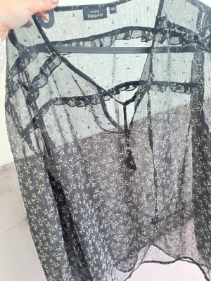 Svart transparent blus från KappAhl - Säljer en svart transparent blus från KappAhl med tunna långärmar och ett diskret blommönster i vitt och gult. Materialet är lätt och luftigt, perfekt för att skapa en cool look. Blusen har en rund halsringning och är riktigt snygg att styla.