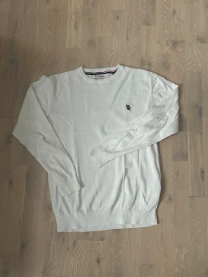 Vit tröja från U.S. Polo Assn - Klassisk vit tröja från U.S. Polo Assn med rund halsringning och diskret logga broderad på bröstet. Tröjan har långa ärmar och ribbade muddar vid ärmslut och nederkant. Perfekt för en clean och stilren look.