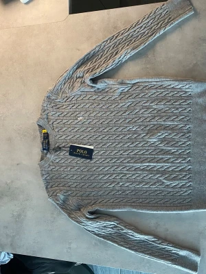 Grå kabelstickad tröja Polo Ralph Lauren - Snygg grå kabelstickad tröja från Polo Ralph Lauren med rund halsringning och diskret broderad logga på bröstet. Tröjan har ribbade muddar vid ärmslut och nederkant. Perfekt för dig som gillar klassisk och stilren design.