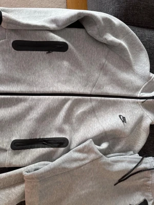 Grå Nike Tech Fleece tracksuit - Snygg grå Nike Tech Fleece hoodie med hel dragkedja och svarta detaljer på fickorna. Endast använd ett par gånger och fortfarande jättefräsch och ren. Storlek M men kan passa L. 