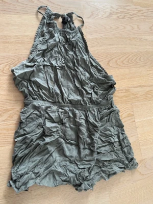 Grågrön kort jumpsuit med halterneck - Superskön grågrön jumpsuit med halterneck och resår i midjan. Tillverkad i mjukt, tunt tyg som känns luftigt och perfekt för varma dagar. Kort modell med avslappnad passform och snygga detaljer vid axelbanden.