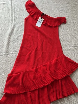 Mng Röd volangklänning från Mango med offshoulder - Snygg röd klänning från Mango med offshoulder och stora volangdetaljer både upptill och nertill. Klänningen har en asymmetrisk nederkant och är perfekt för dig som vill sticka ut. Materialet känns lätt och luftigt, och modellen är feminin och trendig. Nypris 1300kr Passar even S