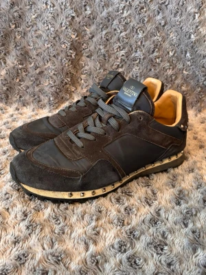Valentino Garavani svarta sneakers - Snygga svarta sneakers från Valentino Garavani med detaljer i mocka och textil. Skorna har grå snören, guldfärgade nitar runt sulan och beige innerfoder. Perfekta för dig som vill ha en lyxig och edgy look.