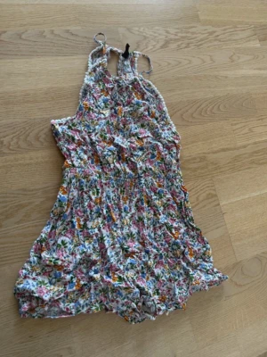 Blommig somrig jumpsuit med smock - Säljer en blommig jumpsuit med smock i midjan och tunna axelband. Tyget är lätt och luftigt, perfekt för varma dagar. Färgerna går i vitt, rosa, blått, grönt och orange. Modellen har korta ben och en avslappnad passform. Inget synligt märke.