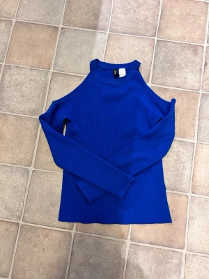 Blå ribbad offshoulder topp från H&M - Snygg blå ribbad topp från H&M med offshoulder-design och långärmade ärmar. Materialet är mjukt och stretchigt, perfekt för att skapa en cool och trendig look. Hög hals och figurnära passform som framhäver axlarna.
