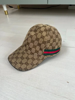Beige Gucci keps med GG-mönster - Säljer en beige keps från Gucci med klassiskt GG-mönster över hela kepsen. På sidan finns en ikonisk grön och röd rand. Kepsen är tillverkad i canvasmaterial och har böjd skärm. Perfekt för dig som vill ha en snygg och trendig accessoar.