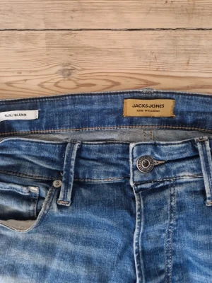 Blå slim fit jeans från Jack & Jones - Snygga blå slim fit jeans från Jack & Jones, modell Glenn. Jeansen har klassisk femficksdesign, normal midja och slitningar för en avslappnad look. Tillverkade i denim med lätt tvättad effekt och kontrastsömmar.