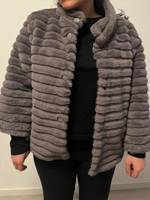 Trekvartsärmad jacka - Säljer en grå pälsjacka med breda horisontella ränder och stora knappar framtill. Jackan har en hög krage och är fodrad med quiltat tyg på insidan. Pälsen är mjuk och syntetisk, perfekt för kalla vinterdagar.