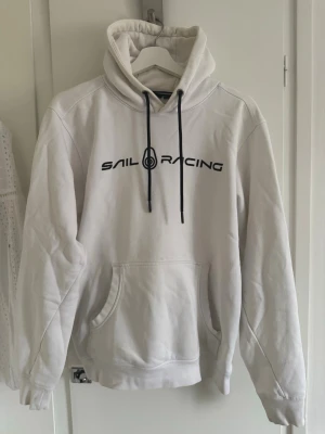 Sail Racing Hoodie - Snygg vit hoodie från Sail Racing med svart logga tryckt på bröstet och en mindre logga på huvan. Har en liten fläck (se bild 3).