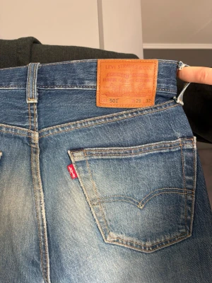 Levi's 501 blå jeans med vintage-look - Klassiska blå Levi's 501 jeans med rak passform och snygg vintage-tvätt. Jeansen har fem fickor, läderpatch bak i midjan och den ikoniska röda Levi's-taggen på bakfickan. Tillverkade i slitstark bomullsdenim och perfekta för en avslappnad stil.