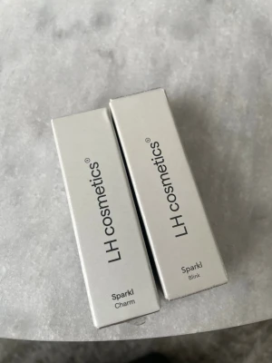 LH cosmetics highlighter Sparkle duo blink charm - Två highlighters från LH cosmetics i nyanserna Sparkle Charm och Sparkle Blink. Förpackningarna är stilrena i silver och guld, och produkterna ger en glittrig finish. Perfekta för att framhäva ansiktets drag med en skimrande look.