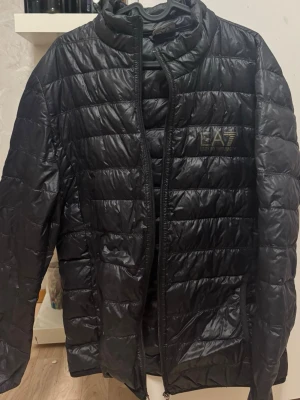 Svart pufferjacka från EA7 Emporio Armani - Snygg svart pufferjacka från EA7 Emporio Armani med quiltad design och hög krage. Jackan har dragkedja framtill och diskret logga på bröstet. Tillverkad i lätt, glansigt material som ger en modern look. Säg till om du behöver fler bilder 🙌