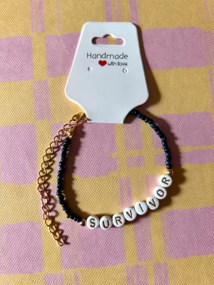 Pärlarmband med texten SURVIVOR - Ett handgjort armband med små svarta pärlor och vita bokstavspärlor som bildar ordet SURVIVOR. Armbandet har guldfärgad kedja och justerbart lås. Perfekt accessoar för att uttrycka styrka och stil.