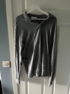 Grå långärmad tröja med krage - En stilren grå långärmad tröja med klassisk krage. Tröjan har en enkel design utan synliga loggor eller mönster och är tillverkad i ett mjukt, tunt material som känns skönt mot huden. Perfekt för dig som gillar minimalistisk stil.