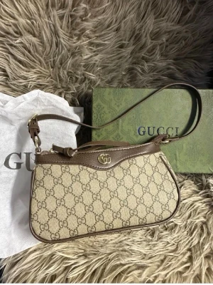 Beige axelremsväska  - Snygg beige handväska med klassiskt GG-monogram i canvas och bruna läderdetaljer. Väskan har en axelrem, guldfärgad logga framtill och dragkedja upptill. Perfekt för dig som gillar lyxiga accessoarer och tidlös design.