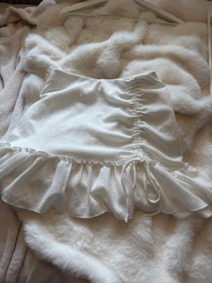 Vit volangkjol från Gina Tricot - Supersöt vit kjol från Gina Tricot med volangkant och snörning på sidan. Kjolen har en kort passform och är gjord i ett mjukt, lätt material som passar perfekt till varmare dagar. Snygg detalj med rynkning framtill och elastisk midja.