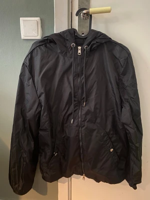 Svart vindjacka från Moncler - Snygg svart vindjacka från Moncler med huva och dragkedja framtill. Jackan har två fickor med knapp, justerbar dragsko i huvan och Moncler-logga på ärmen. På huvan finns en röd och vit rand som detalj. Tillverkad i lätt och tåligt material.