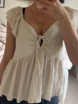 Beige omlottopp med volangärm - Söt beige topp med omlottdesign och knyt framtill. Toppen har volangärmar och en lätt rynkad, luftig passform som ger en romantisk känsla. Perfekt till jeans eller kjol för en trendig look.