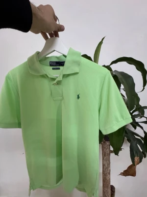 Grön pikétröja från Polo Ralph Lauren - Fräsch ljusgrön pikétröja från Polo Ralph Lauren med klassisk krage och korta ärmar. Tröjan har den ikoniska broderade loggan på bröstet och är tillverkad i mjuk bomull. Perfekt för en avslappnad och stilren look.