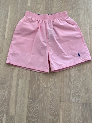 Rosa shorts från Polo Ralph Lauren - Snygga ljusrosa shorts från Polo Ralph Lauren med elastisk midja och klassisk blå logga broderad på ena benet. Perfekta för sommaren och tillverkade i ett lätt och bekvämt material.