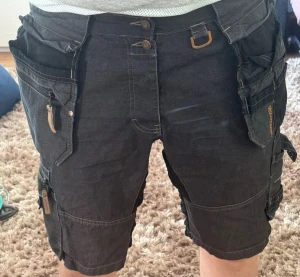 Svarta cargoshorts från SOUL - Tuffa svarta cargoshorts från SOUL med flera stora fickor, dragkedjor och detaljer i brons. Shorts med normal passform och praktiska fickor på sidorna, perfekta för dig som gillar funktionella plagg. Materialet är bomull och polyester.