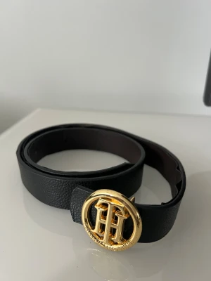 Svart skärp med guldspänne Tommy Hilfiger - Stilrent svart skärp från Tommy Hilfiger med ett rundat guldspänne som har TH-loggan. Skärpet är tillverkat i skinn och har en klassisk, enkel design som passar till jeans eller kostymbyxor. Perfekt för att lyfta din outfit med en touch av lyx. Se skick på bild 