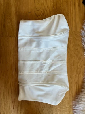 Vit satängbody med bandeauform - Snygg vit body i glansigt satängmaterial med bandeauform och markerade sömmar framtill. Perfekt för dig som gillar en stilren och elegant look. Materialet är mjukt och följsamt mot huden.