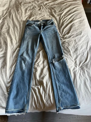 Blå raka jeans med slitningar - Säljer ett par blå raka jeans med slitningar på ena benet och fransade benslut. Jeansen har klassisk femficksdesign och är i en ljusblå tvätt. Perfekta för en avslappnad och trendig look. Jag har sprättat upp de nertill💕långa på mig som är 175+