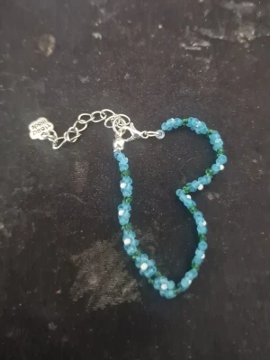 Blått pärlarmband med blommor - Ett handgjort armband med små blå och gröna pärlor formade som blommor. Armbandet har en silverfärgad kedja och justerbart lås. Perfekt accessoar för att ge din outfit en färgklick och en söt detalj.