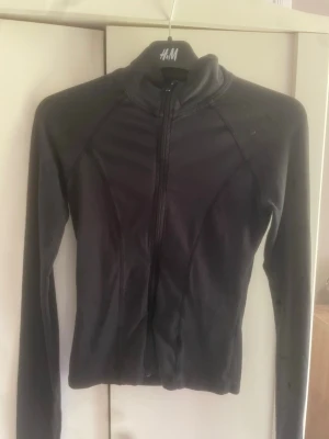 Svart sportjacka med dragkedja - Svart sportjacka från gina  med hel dragkedja framtill och lång ärm. Jackan har en figurnära passform och är tillverkad i ett stretchigt, lätt material som passar perfekt till träning eller löpning. ett litet håll men inget som märks när man har på sig den.