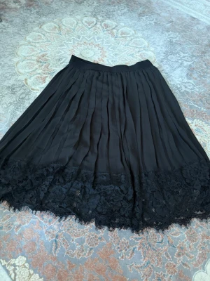 Svart plisserad kjol med spetskant - Svart plisserad kjol med bred spetskant nertill. Kjolen har en flowy känsla och är perfekt för dig som gillar en klassisk men ändå trendig look. Resår i midjan för extra komfort och snygg passform.