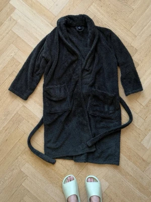 Mysig mörkgrå fleece morgonrock - Supermjuk och varm morgonrock i mörkgrå fleece med långa ärmar, två stora fickor framtill och knytskärp i midjan. Perfekt för chilliga morgnar eller kvällar hemma. Enkel och stilren design utan synliga loggor.