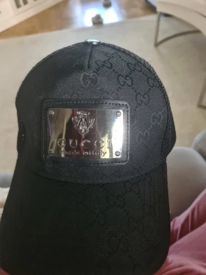 Svart keps från Gucci med metallbricka - Svart keps från Gucci med GG-monogram över hela tyget och en snygg metallbricka framtill med logga. Kepsen är tillverkad i bomull och polyester med lite elastan för stretch. Perfekt för dig som vill ha en lyxig och trendig accessoar.