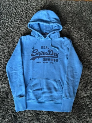 Vintage Superdry hoodie - Small - Superdry hoodie i vintage stil i storlek S. Hoodien har en stilren blå färg med klassiskt Superdry-tryck framtill som ger en tidlös och snygg look. Tillverkad i ett mjukt och bekvämt material som passar perfekt till vardags.  Använd endast fåtal gånger och är därför i mycket fint skick, nästan som ny.   Storlek S. Se sista bilderna för mått🙂 Nästan ny, använd fåtal ggr.  Fråga vid funderingar:) Skickas snabbt📦 