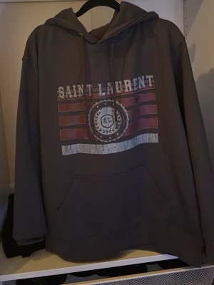 Saint Laurent brun hoodie med tryck - Cool grå hoodie från Saint Laurent med tryck framtill i vitt och rött. Klassisk huva med dragsko och stor magficka. Perfekt för en avslappnad streetstyle-look.