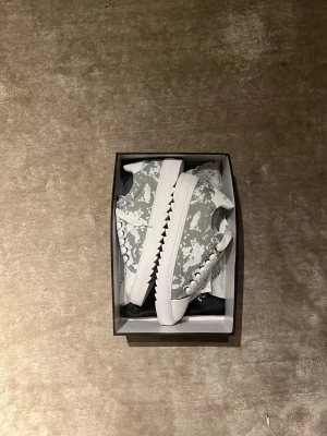Gråvita sneakers från Numeris - Snygga sneakers från Numeris med gråvitt camomönster och vita detaljer. Skorna har tjock vit sula med taggad kant och breda vita skosnören. Ovandelen är i textil och läderimitation. Perfekta för dig som vill sticka ut med en unik look.